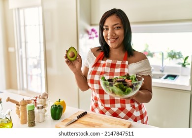 Hispanic Brunette Woman Preparing Green Salad Holding Avocado At The Kitchen