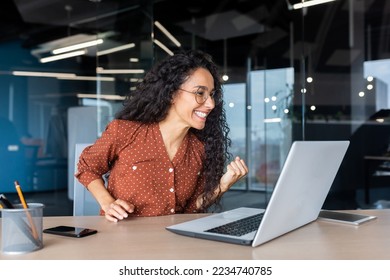 Hispanic business woman celebrating victory success, employee with curly hair inside office reading good news, using laptop at work inside office holding hand up and happy triumph gesture.