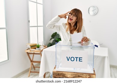 Hispanic business woman voting putting envelop in ballot box doing peace symbol with fingers over face, smiling cheerful showing victory 