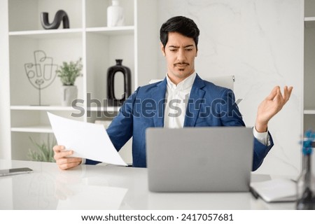 Hispanic businessman in a blue suit expressing confusion or concern while holding a document, seated at his office desk with a laptop, depicting a moment of business challenge or problem-solving