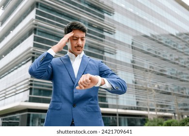 A Hispanic businessman in a blue suit looks concerned while checking the time on his watch outside a corporate building, representing the fast-paced nature of business, feels leak of time and deadline - Powered by Shutterstock