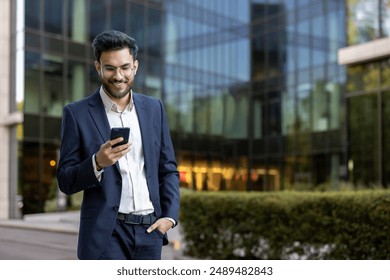 Hispanic businessman in formal attire using smartphone outside modern office building. Man smiling, engaging with technology, and exuding confidence. Corporate environment glass facade in background. - Powered by Shutterstock