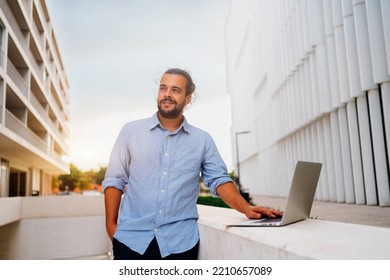 Hispanic Businessman Standing Outside With Laptop Working Remote Distant Job Outdoors Near Office Company Building. Stylish Unshaved Male Business Man With Long Hair Dressed Use Laptop