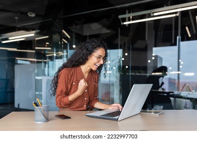 Hispanic businesswoman celebrating victory and successful achievement, office worker received online news and happy smiling and looking at laptop screen holding hand up triumph gesture. - Shutterstock ID 2232997703