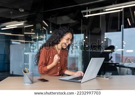 Hispanic businesswoman celebrating victory and successful achievement, office worker received online news and happy smiling and looking at laptop screen holding hand up triumph gesture.