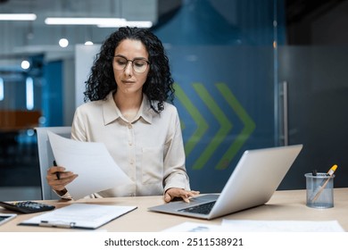 Hispanic businesswoman engaged in office work, reviewing important documents and using laptop. Professional setting depicts business environment, showcasing focus and management skills. - Powered by Shutterstock