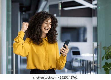 Hispanic businesswoman exudes excitement and triumph checking phone in office. Curly hair, bright yellow blouse, fist pump conveying victory. Modern work setting highlights professional atmosphere - Powered by Shutterstock