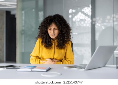 Hispanic businesswoman sits at office desk, experiencing stomach pain. She holds her abdomen, showing stress and discomfort. Nearby are laptop and notebook, indicating a work setting. - Powered by Shutterstock