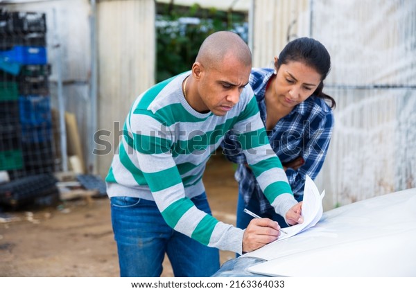 Hispanic couple of farmers\
discussing some papers while standing near car on backyard of\
farmstead