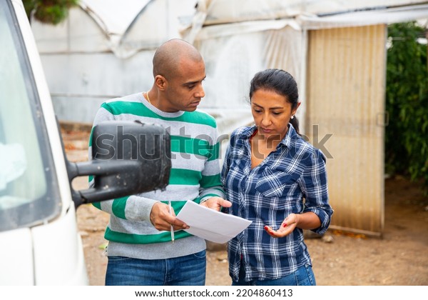 Hispanic couple of farmers\
discussing some papers while standing near car on backyard of\
farmstead