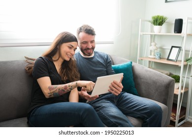 Hispanic Couple Looking At Social Media On A Tablet And Resting On The Couch On A Leisure Day At Home. Happy Husband And Wife Hugging While Doing Online Shopping