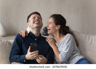 Hispanic Couple Sit On Sofa In Living Room With Smart Phone, Laughing Over Online Videos, Watch Prank On Internet Looking Overjoyed Spend Time At Home Use Virtual Amusement And Entertainment Concept