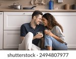 Hispanic couple sit on warm floor with mugs in cozy kitchen, enjoy romantic dating, house-warming, pleasant talk and beverage, spend relocation day to new own house. Cohabitation, relationships, love