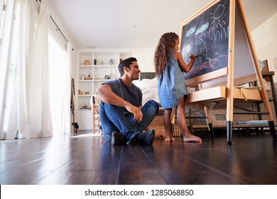 Hispanic Dad Sitting On The Floor In Sitting Room Watching His Young Daughter Drawing On Blackboard