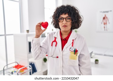 Hispanic Doctor Woman With Curly Hair Holding Heart Thinking Attitude And Sober Expression Looking Self Confident 