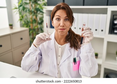 Hispanic Doctor Woman Holding Blood Sample And Syringe Puffing Cheeks With Funny Face. Mouth Inflated With Air, Catching Air. 