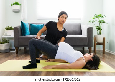 Hispanic doula helping an attractive pregnant woman to do a bridge yoga pose. Expectant mother working out with her midwife in the living room