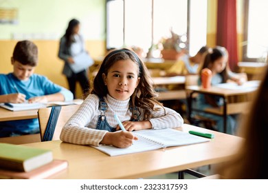 Hispanic elementary student writing in her notebook on a class at school and looking at camera. - Shutterstock ID 2323317593