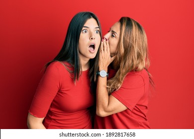 Hispanic family of mother and daughter wearing casual clothes over red background hand on mouth telling secret rumor, whispering malicious talk conversation 