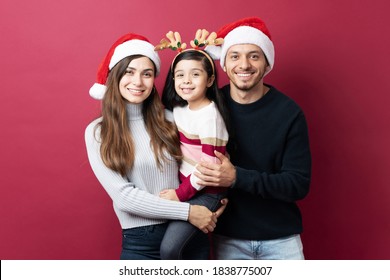 Hispanic Family Of Three Hugging Each Other Standing Against A Red Background On Christmas