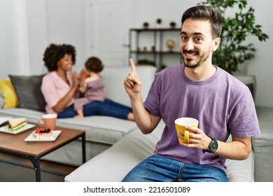 Hispanic Father Of Interracial Family Drinking A Cup Coffee With A Big Smile On Face, Pointing With Hand Finger To The Side Looking At The Camera. 