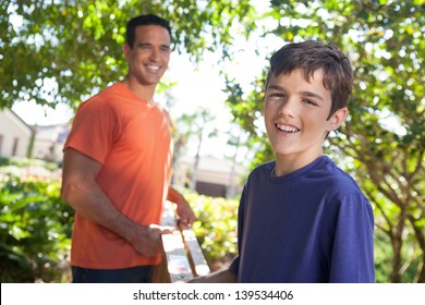 Hispanic Father And Teenaged Son Happily Carry Ladder Together Outside In Yard.