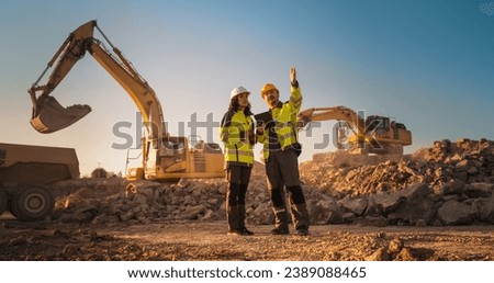 Hispanic Female Inspector Talking to Caucasian Male Land Development Manager With Tablet On Construction Site Of Real Estate Project. Excavators Preparing For Laying Building Foundation. Hot Sunny Day