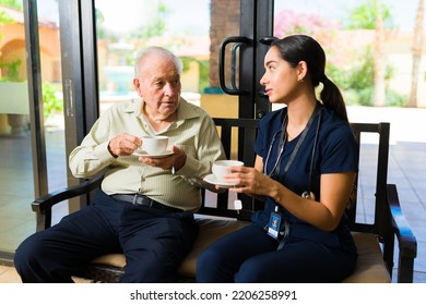 Hispanic Female Nurse Working At A Retirment Home Drinking A Cup Of Coffee While Chatting With A Retired Mature Man 