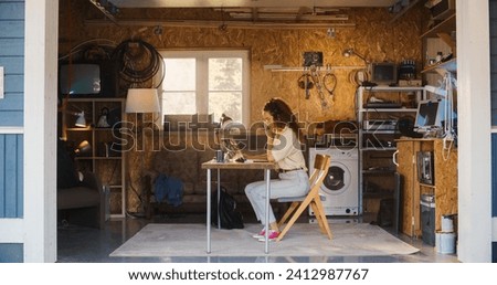 Hispanic Female Software Engineer Programming On Laptop Computer In Garage At Home. Focused Young Woman Working On Innovative Online Service Startup Company, Coding. Digital Entrepreneur Concept.