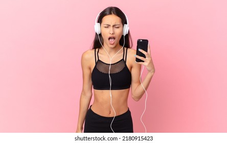 Hispanic Fitness Woman Shouting Aggressively, Looking Very Angry With Headphones And A Phone