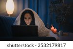 Hispanic freelancer girl Latino woman Indian Arabian female at night evening home at couch sofa under blanket cover duvet working with laptop watching movie at computer browsing social media addict