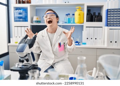 Hispanic girl with down syndrome working at scientist laboratory crazy and mad shouting and yelling with aggressive expression and arms raised. frustration concept.  - Shutterstock ID 2258990131