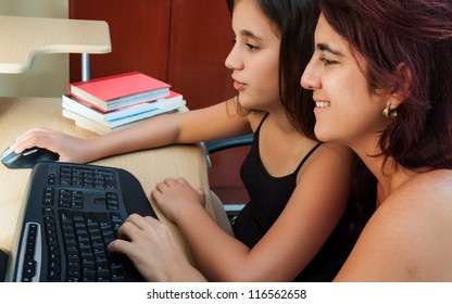 Hispanic Girl And Her Young Mother Using A Computer At Home Wit Books On The Computer Desk