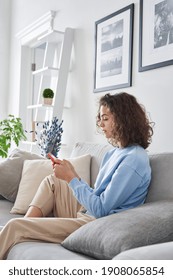 Hispanic Girl Holding Cell Phone Sitting On Sofa At Home. Young Latin Woman Using Smartphone Mobile Apps On Cellphone Device In Apartment, Teen Tech Addiction, Information Overload Concept.