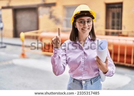 Hispanic girl wearing architect hardhat at construction site smiling with an idea or question pointing finger with happy face, number one 