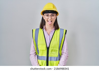 Hispanic girl wearing builder uniform and hardhat sticking tongue out happy with funny expression. emotion concept. 