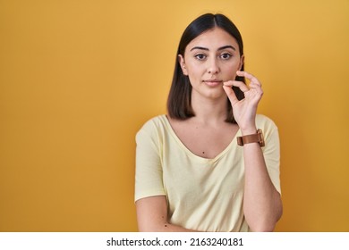 Hispanic girl wearing casual t shirt over yellow background mouth and lips shut as zip with fingers. secret and silent, taboo talking 