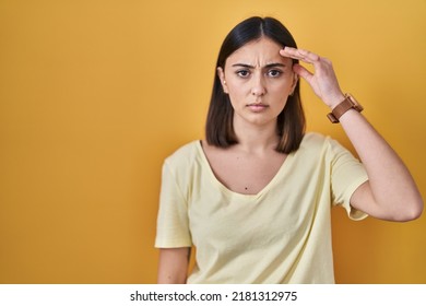 Hispanic girl wearing casual t shirt over yellow background pointing unhappy to pimple on forehead, ugly infection of blackhead. acne and skin problem 