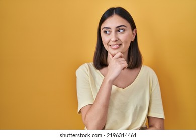 Hispanic Girl Wearing Casual T Shirt Over Yellow Background With Hand On Chin Thinking About Question, Pensive Expression. Smiling And Thoughtful Face. Doubt Concept. 