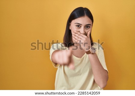 Hispanic girl wearing casual t shirt over yellow background laughing at you, pointing finger to the camera with hand over mouth, shame expression 