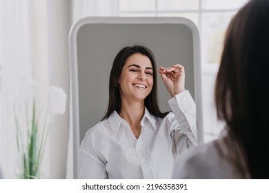 Hispanic Gorgeous Young Woman In White Shirt At Home Looking At Mirror, Toothy Smiling, Satisfied After Cosmetology Procedure, Skin Care, Health And Beauty Concept. Women Health And Beauty.