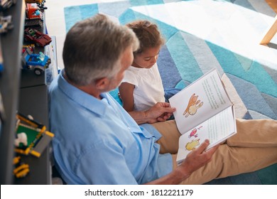 Hispanic Grandfather And Granddaughter Sitting On Floor Of Childrens Bedroom Reading Book Together