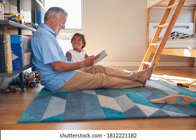 Hispanic Grandfather And Granddaughter Sitting On Floor Of Childrens Bedroom Reading Book Together