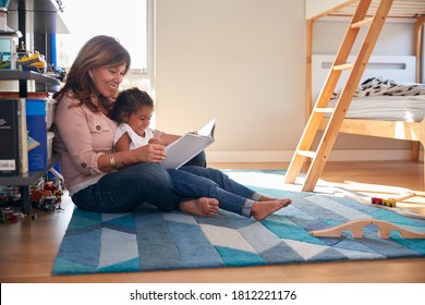 Hispanic Grandmother And Granddaughter Sitting On Floor Of Childrens Bedroom Reading Book Together