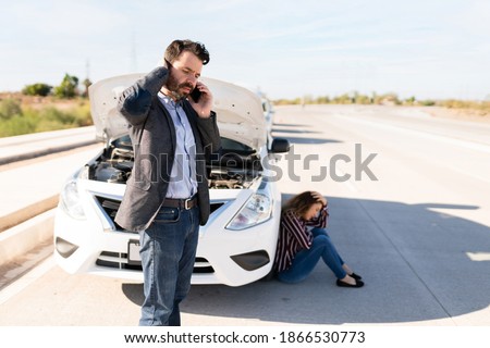 Hispanic guy talking on the phone to ask for a mechanic to come fix their broken-down car in the highway. Sad young woman sitting and waiting for help