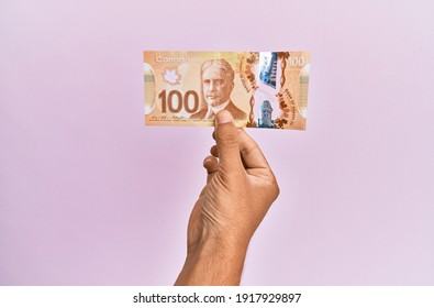 hispanic hand holding 100 canadian dollars banknote over isolated pink background.