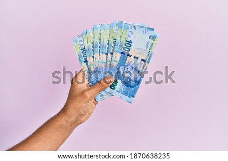 Hispanic hand holding 100 south africa rands banknotes over isolated pink background.