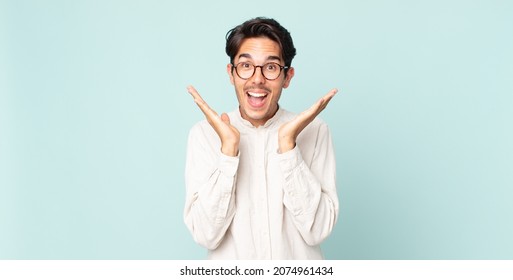 hispanic handsome man looking happy and excited, shocked with an unexpected surprise with both hands open next to face