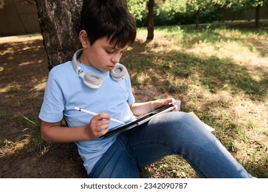 Hispanic handsome smart teenage school boy, using a stylus pen drawing, sketching on a digital tablet while resting outdoors. Children. People. Creativity. Education. Leisure activity. Lifestyle - Powered by Shutterstock