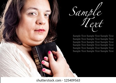 Hispanic Looking Woman In A Light Dress Against A Black Background Holding A Bible Close To Her Body As She Looks Up In Thought. Sample Text To Show How Your Text Can Be Used.